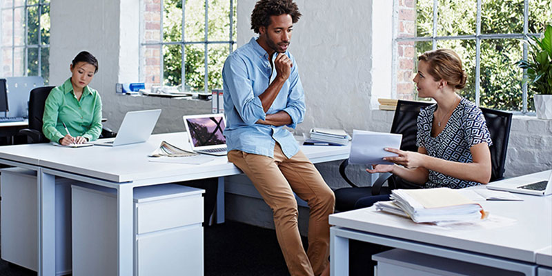 Three co-workers in a brightly lit modern office, with large windows with green plants outside