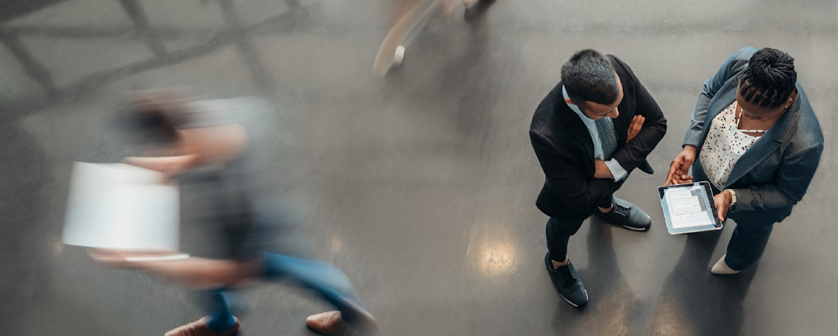 Overhead view of two business people looking at a tablet in a fast paced office. The other people in the image are blurred.