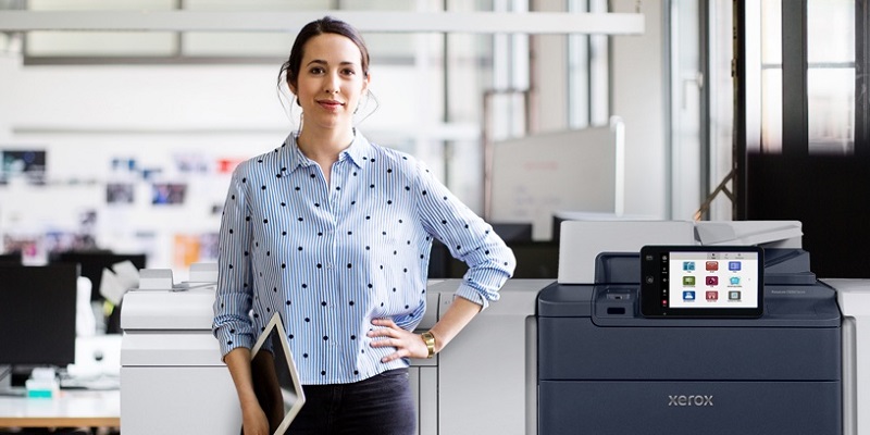 A woman with a tablet in an office, standing next to a Xerox PrimeLink C9200 Series Printer