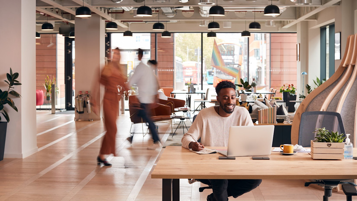 Man sitting in an office on a laptop with people behind him.