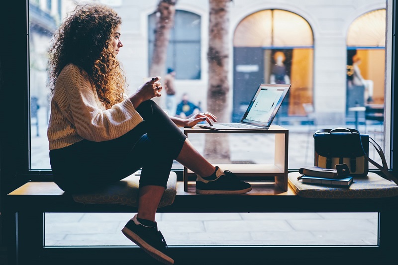 Woman sitting on a table at a window, working on a laptop