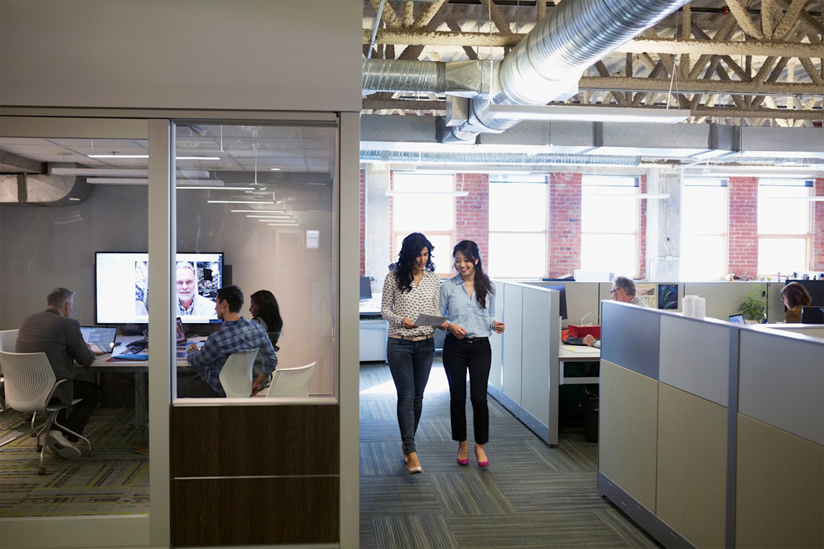 Women walking down an office corridor, talking and looking at printed documents.
