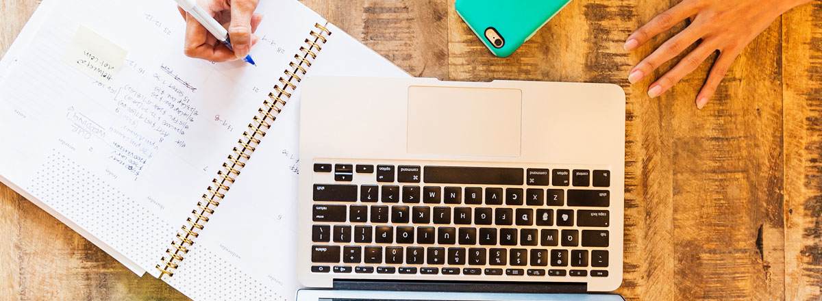 Top view of a desk with a person writing in a notebook, next to a laptop and smarlphone